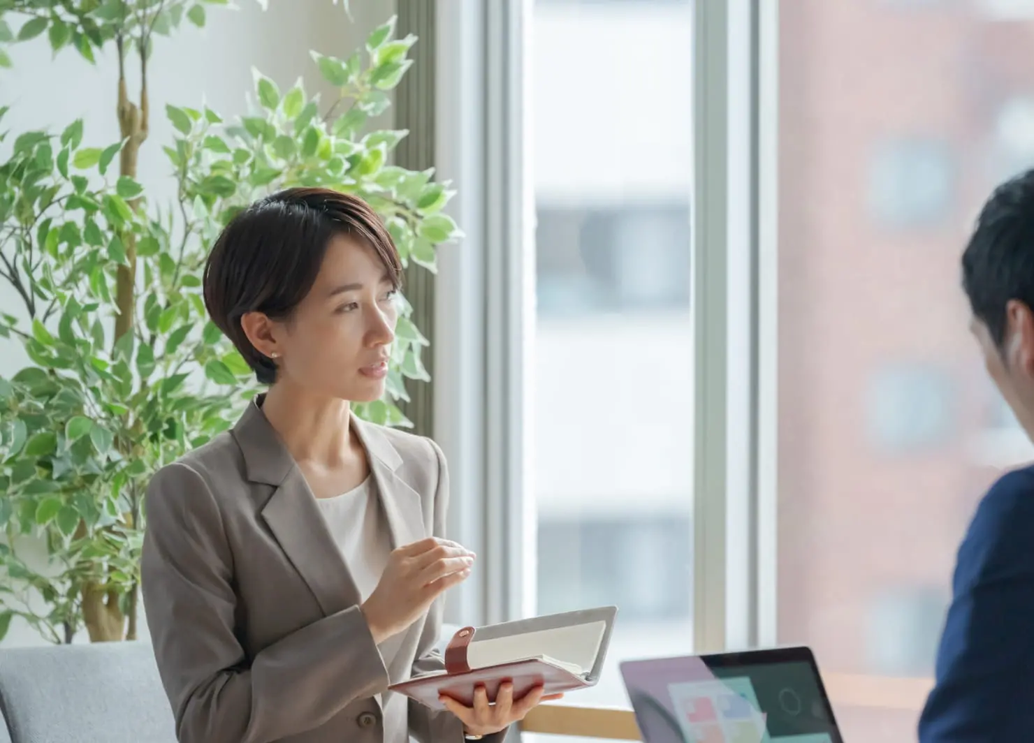 オフィスでメモを手に話をするスーツ姿の女性。対面には同僚が座っている