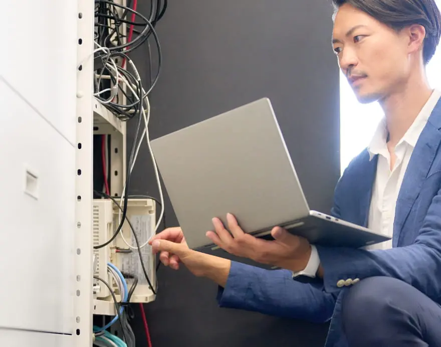 サーバールームでノートパソコンを操作しながら配線を点検する男性の写真