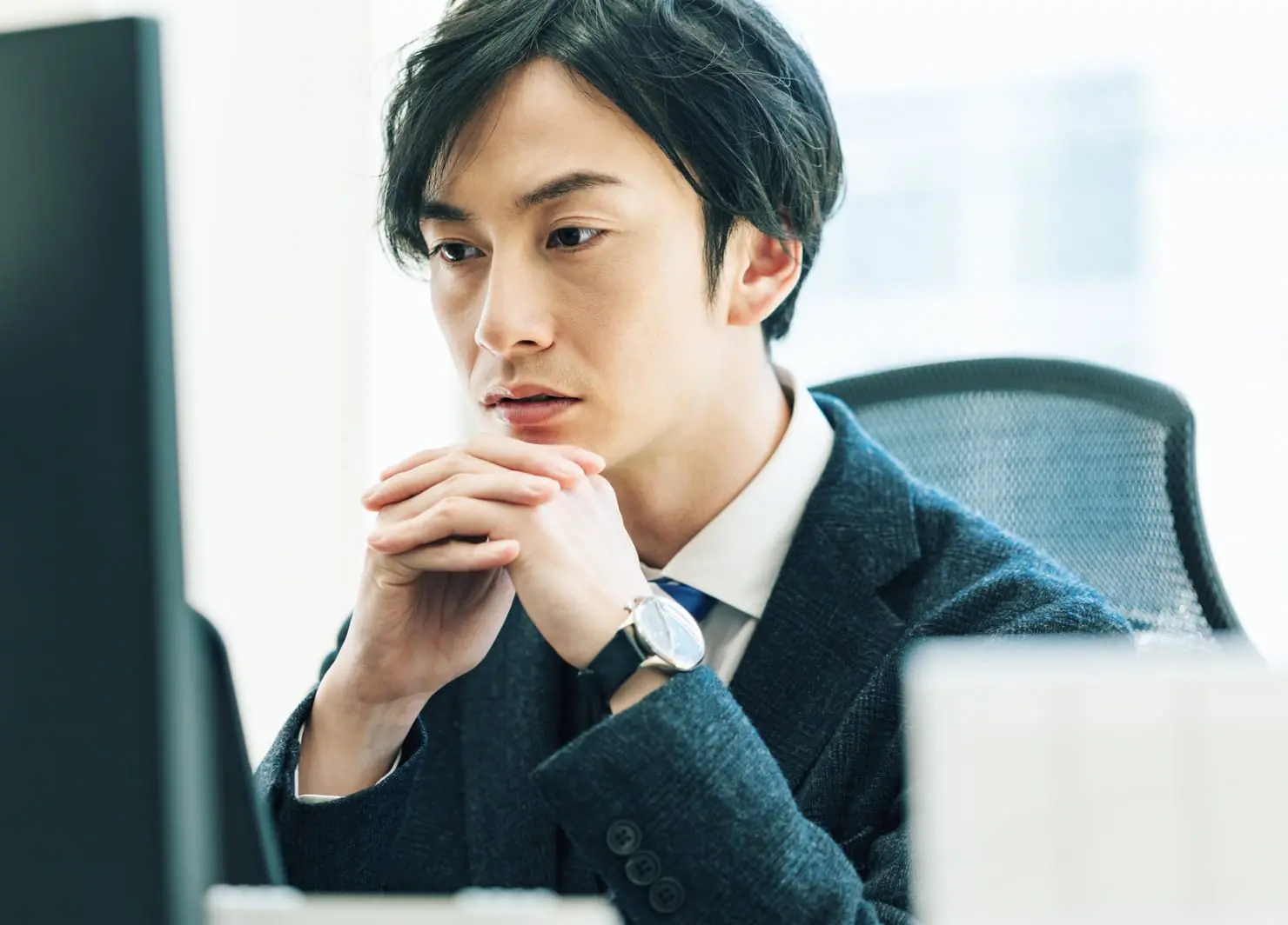 デスクで真剣な表情でモニターを見るスーツ姿の男性の写真