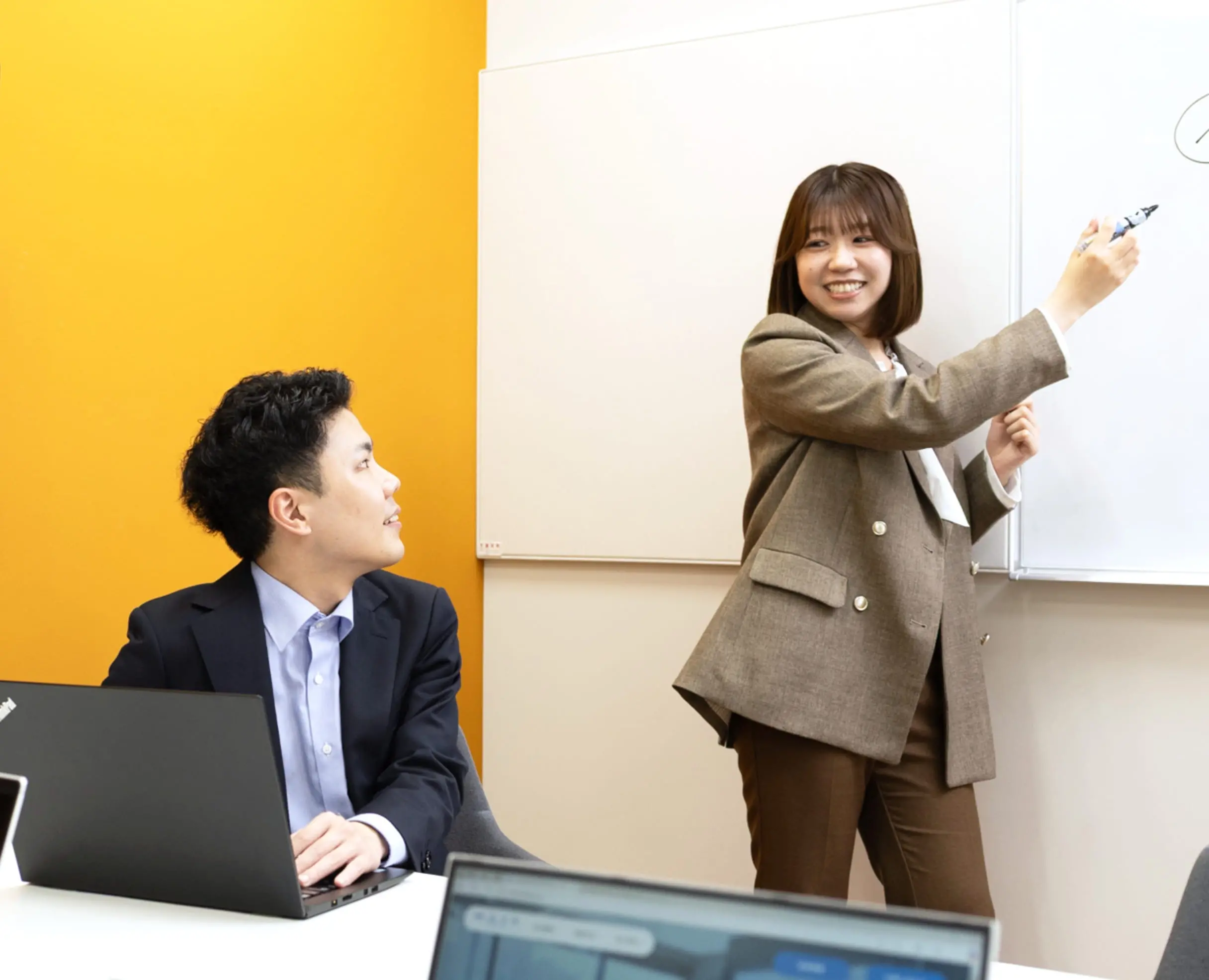 オフィスでホワイトボードを使いながら説明する女性社員と、それを聞く男性社員の写真