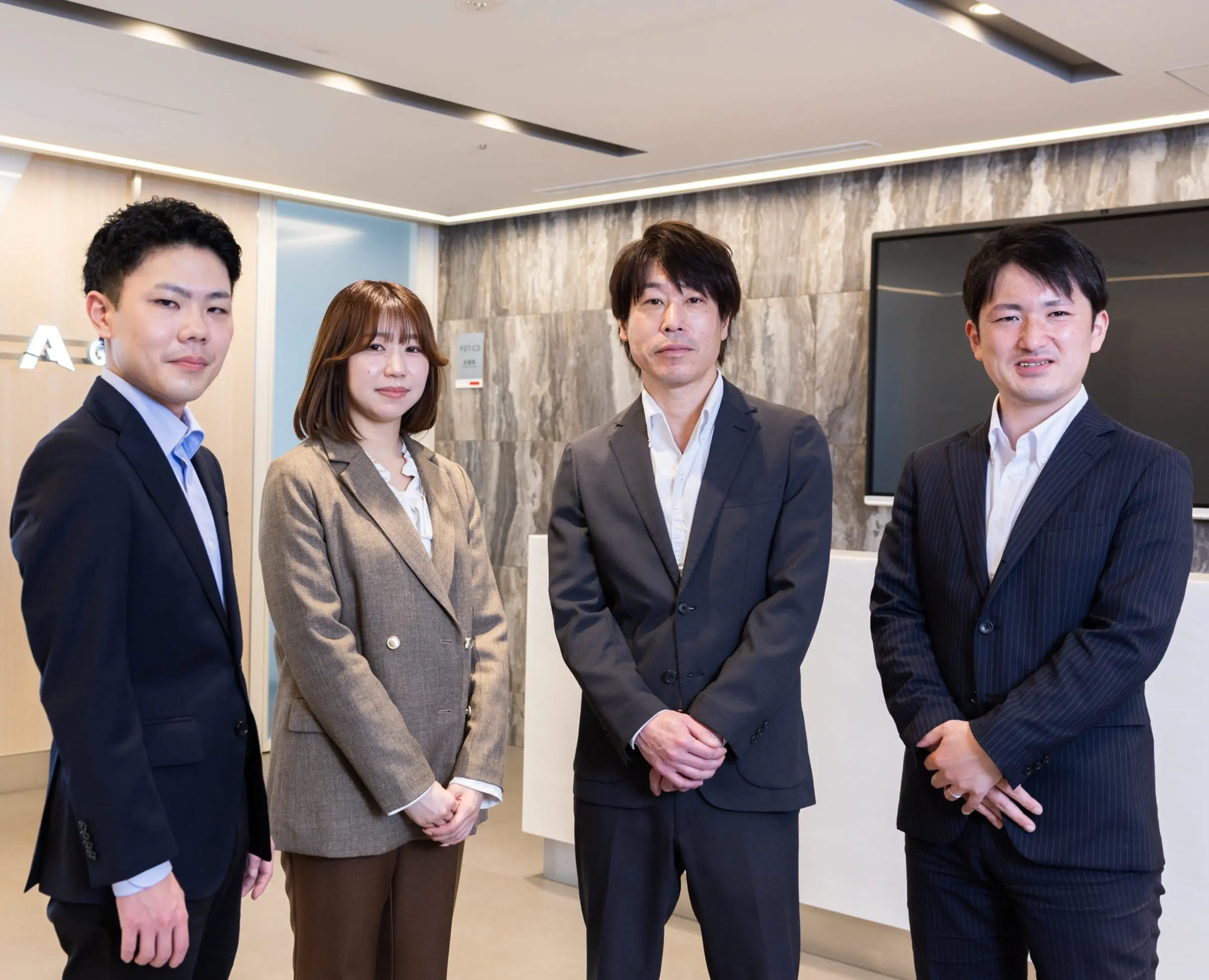 オフィスの受付で並ぶ4人の社員。スーツ姿でカメラを見ている写真